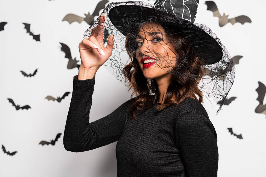 mulher atraente em chapéu de bruxa sorrindo e olhando para a câmera no Halloween
 - Foto, Imagem