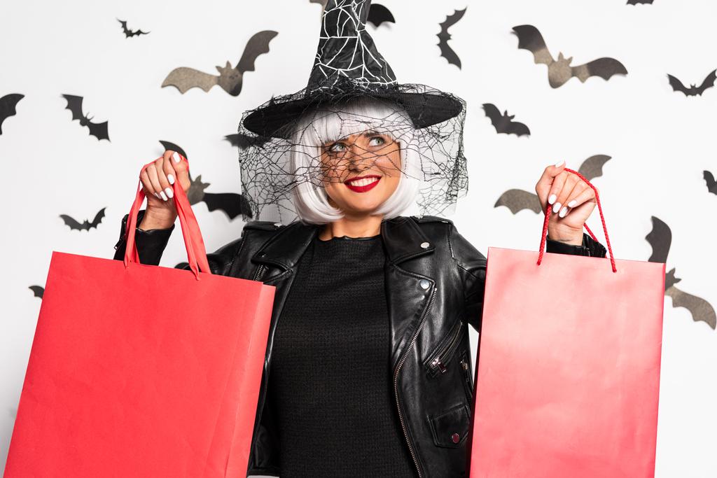 uśmiechnięta kobieta w kapeluszu czarownicy i peruce trzyma torby na zakupy w Halloween - Zdjęcie, obraz