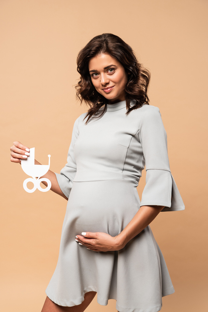 pregnant woman in grey dress holding paper baby carriage on beige background  - Photo, Image