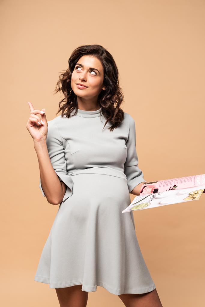 Schwangere im grauen Kleid hält Magazin in der Hand und zeigt Ideengeste auf beigem Hintergrund  - Foto, Bild