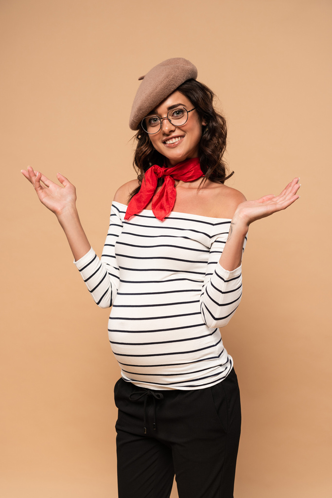 妊娠中のフランス人女性でベレー帽に手を伸ばしてベージュの背景で笑顔 ロイヤリティフリー写真 画像素材