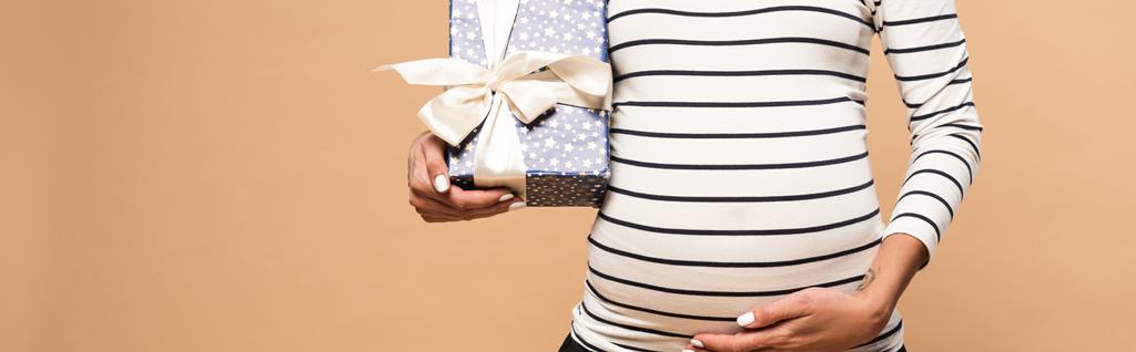 abgeschnittene Ansicht einer schwangeren Frau mit Geschenk auf beigem Hintergrund  - Foto, Bild