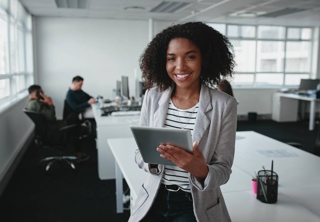 Ritratto di una giovane donna d'affari afro-americana sorridente e sicura di sé che tiene in mano il tablet digitale guardando la macchina fotografica con un collega in background in ufficio
 - Foto, immagini