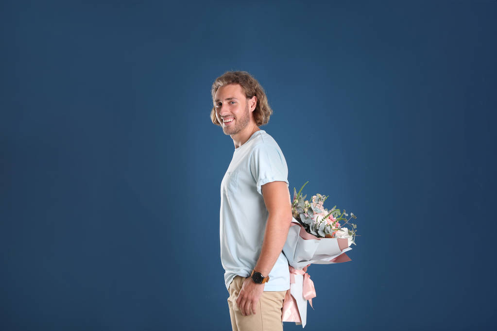Mladý pohledný muž skrývající krásnou květinové Buket za zády na modrém pozadí - Fotografie, Obrázek
