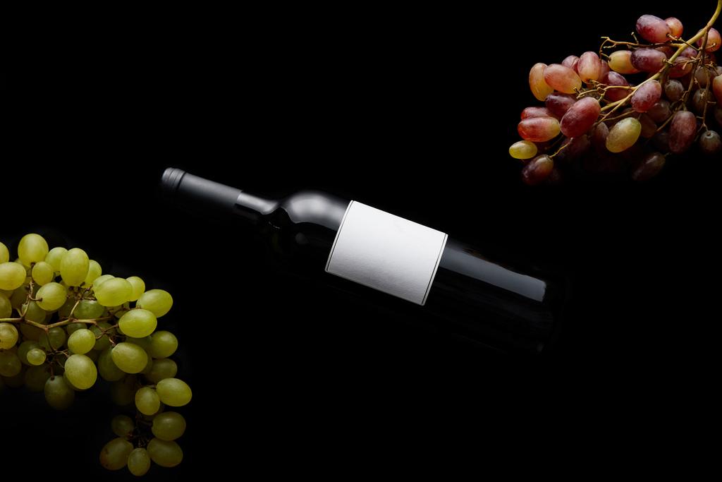 vista dall'alto di bottiglia di vino con etichetta bianca bianca vicino all'uva matura isolata su nero
 - Foto, immagini