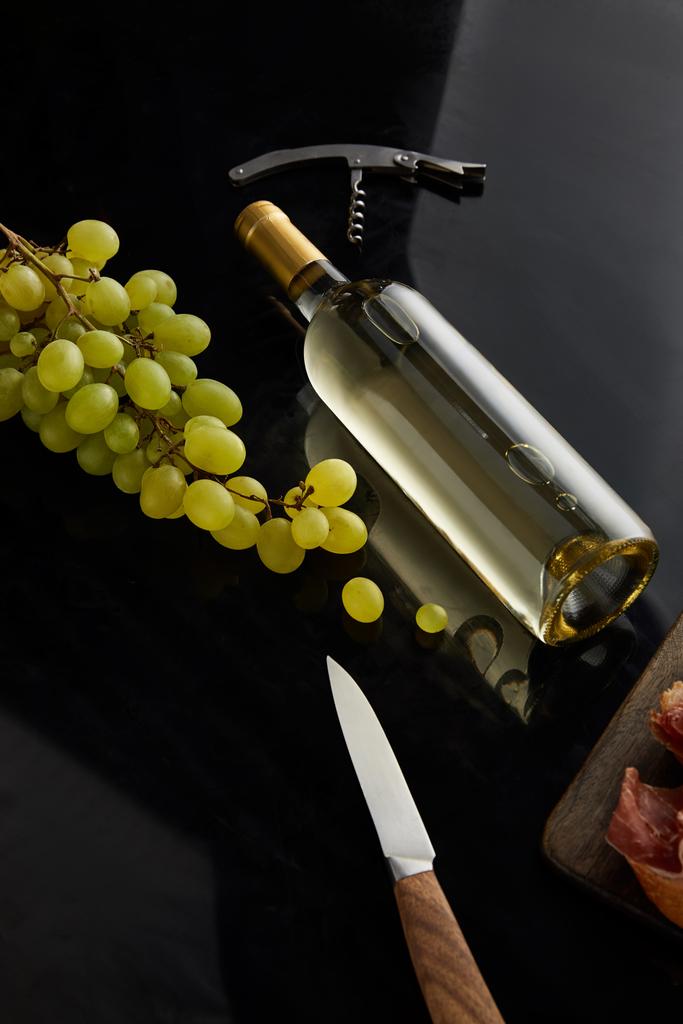 garrafa com vinho branco perto de uva, faca e saca-rolhas na superfície preta
 - Foto, Imagem