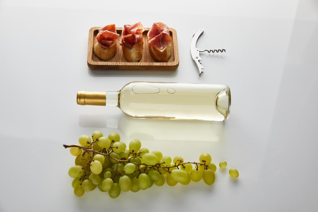 plochý ležel s lahví s bílým vínem v blízkosti hroznů, vývrtky a krájené prosciutto na bagetě na bílém pozadí - Fotografie, Obrázek