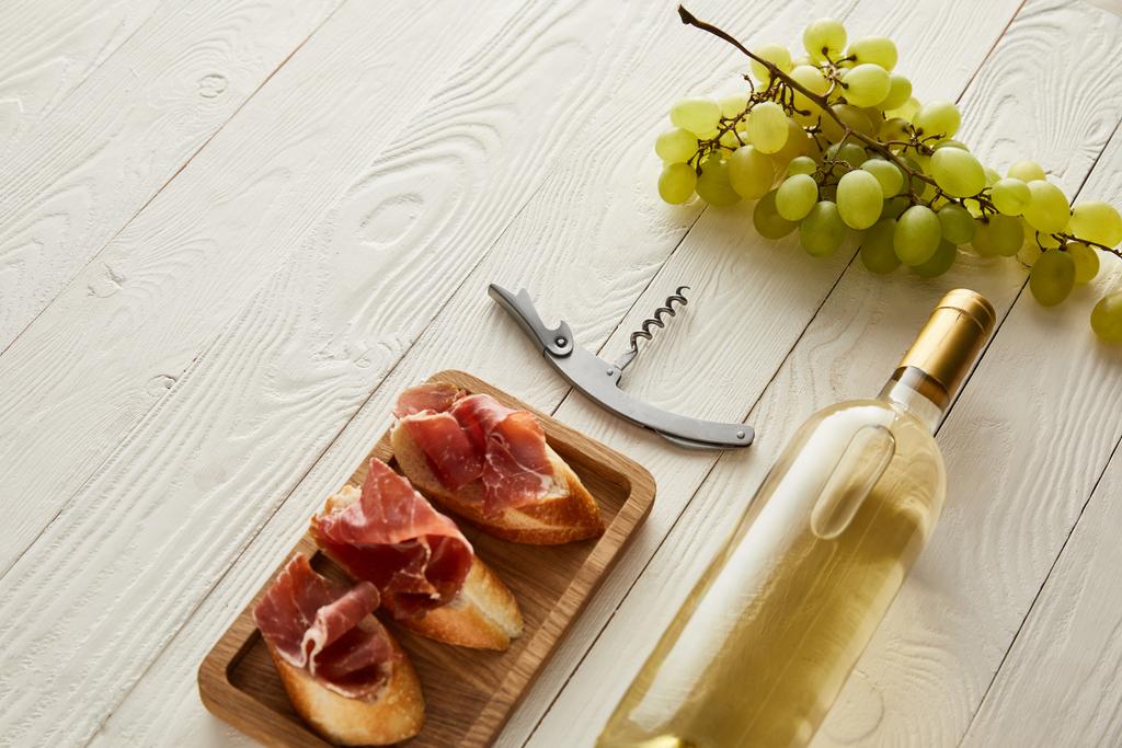 bottle with white wine near grape, prosciutto on baguette and corkscrew on white wooden surface - Photo, Image