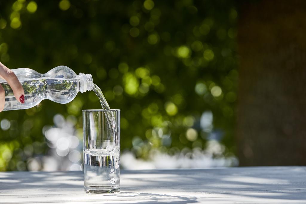 καλλιεργημένη άποψη της γυναίκας ρίχνει φρέσκο καθαρό νερό από το μπουκάλι σε διαφανές γυαλί την ηλιόλουστη μέρα σε ξύλινο τραπέζι - Φωτογραφία, εικόνα