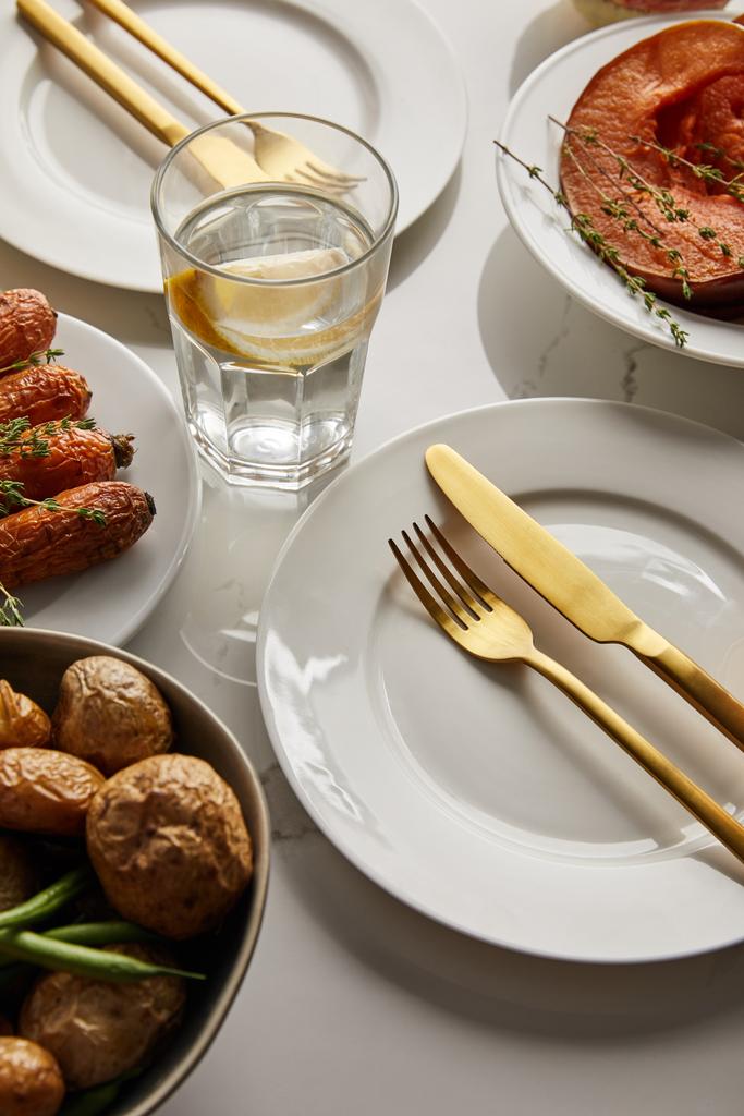 white plates with baked vegetables, golden forks and knives, glass with lemon water on white marble table - Photo, Image