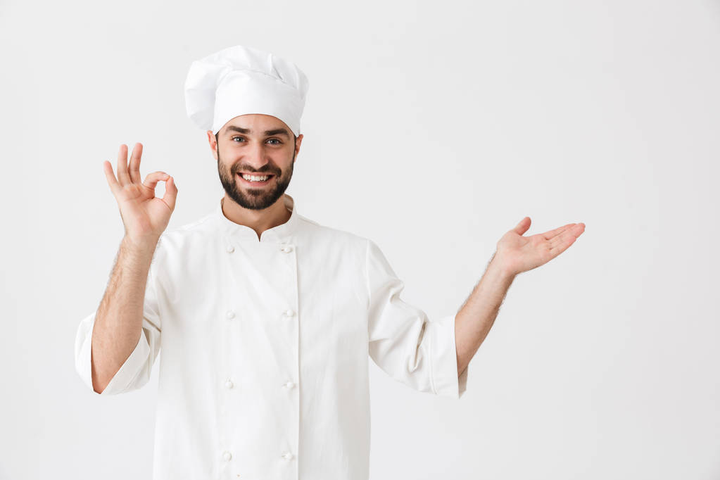 Счастливый молодой шеф-повар позирует изолированно на белом фоне стены в униформе сделать нормальный жест показывая подражания
. - Фото, изображение