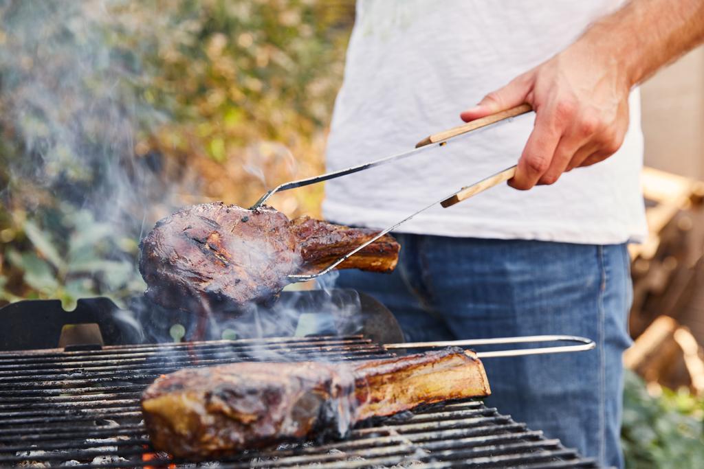 vista ritagliata dell'uomo con pinzette grigliate di carne sulla griglia del barbecue
  - Foto, immagini