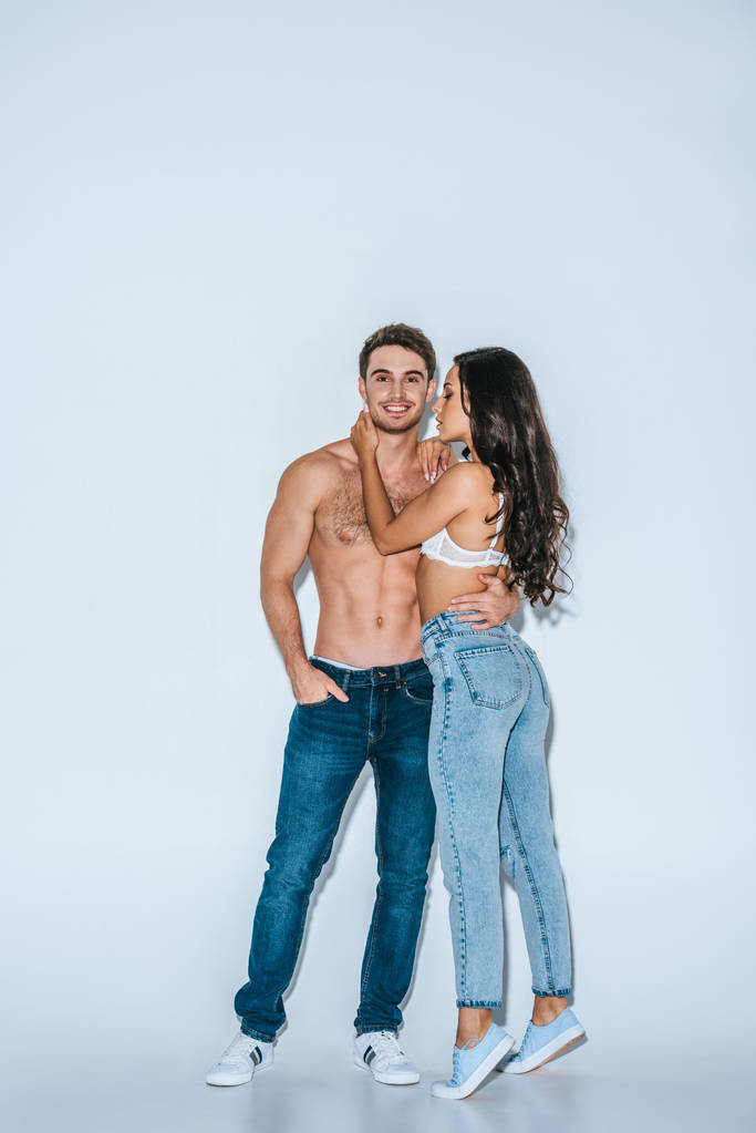 shirtless young man smiling at camera while embracing sexy girl on grey background - Photo, Image