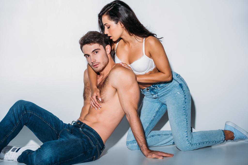 sexy chica en jeans en blanco sujetador abrazando novio sentado y mirando a la cámara en gris fondo
 - Foto, Imagen