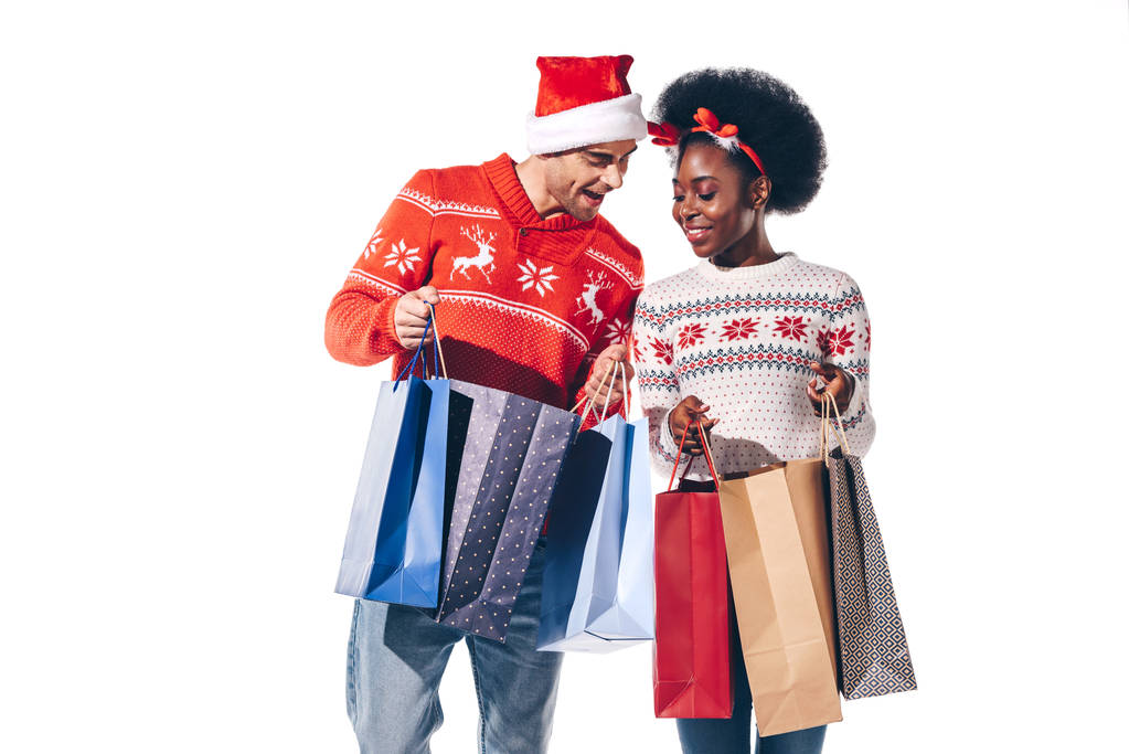 glückliches gemischtrassiges Paar mit Weihnachtsmann und Hirschhörnern in Einkaufstaschen, isoliert auf weißem Grund - Foto, Bild