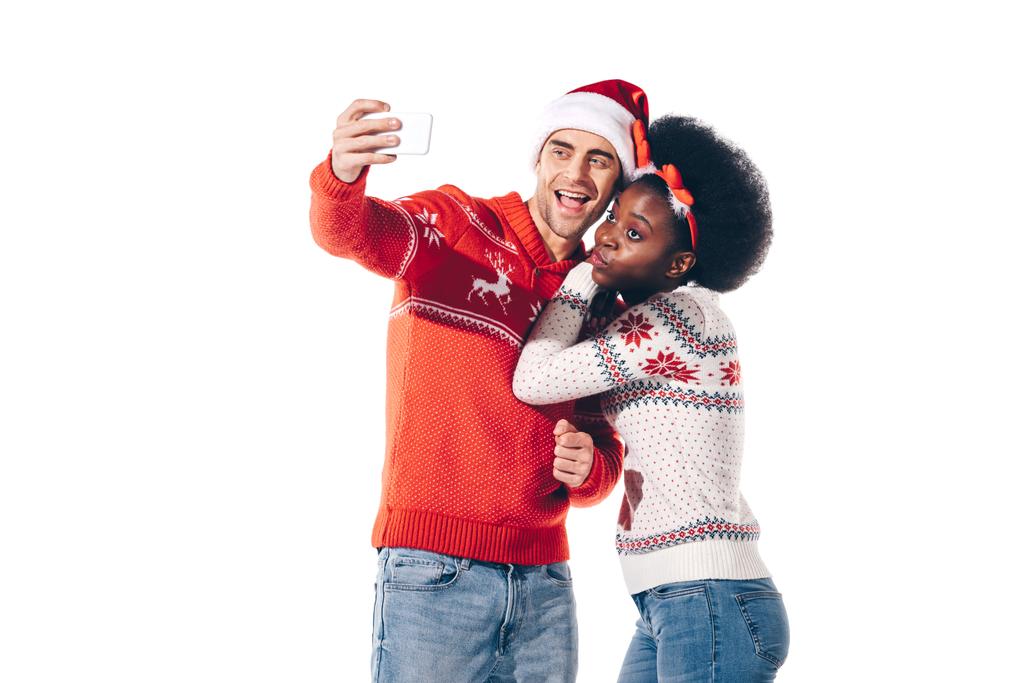 glückliches gemischtrassiges Paar mit Weihnachtsmann und Hirschhörnern, das ein Selfie auf dem Smartphone macht, isoliert auf weiß - Foto, Bild