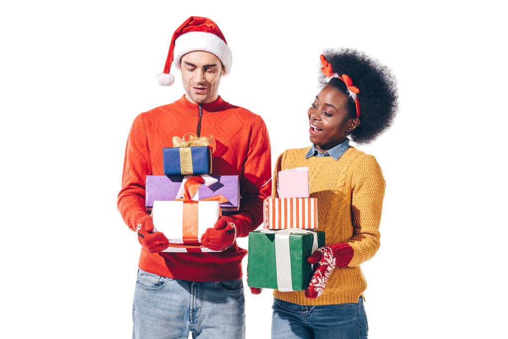 ευτυχισμένο διαφυλετικό ζευγάρι σε καπέλο Σάντα και κέρατα ελαφιού κρατώντας χριστουγεννιάτικα δώρα, απομονωμένο σε λευκό - Φωτογραφία, εικόνα
