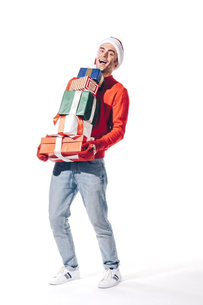 szczęśliwy człowiek w kapeluszu Mikołaja trzymając stos prezentów świątecznych, odizolowany na białym - Zdjęcie, obraz