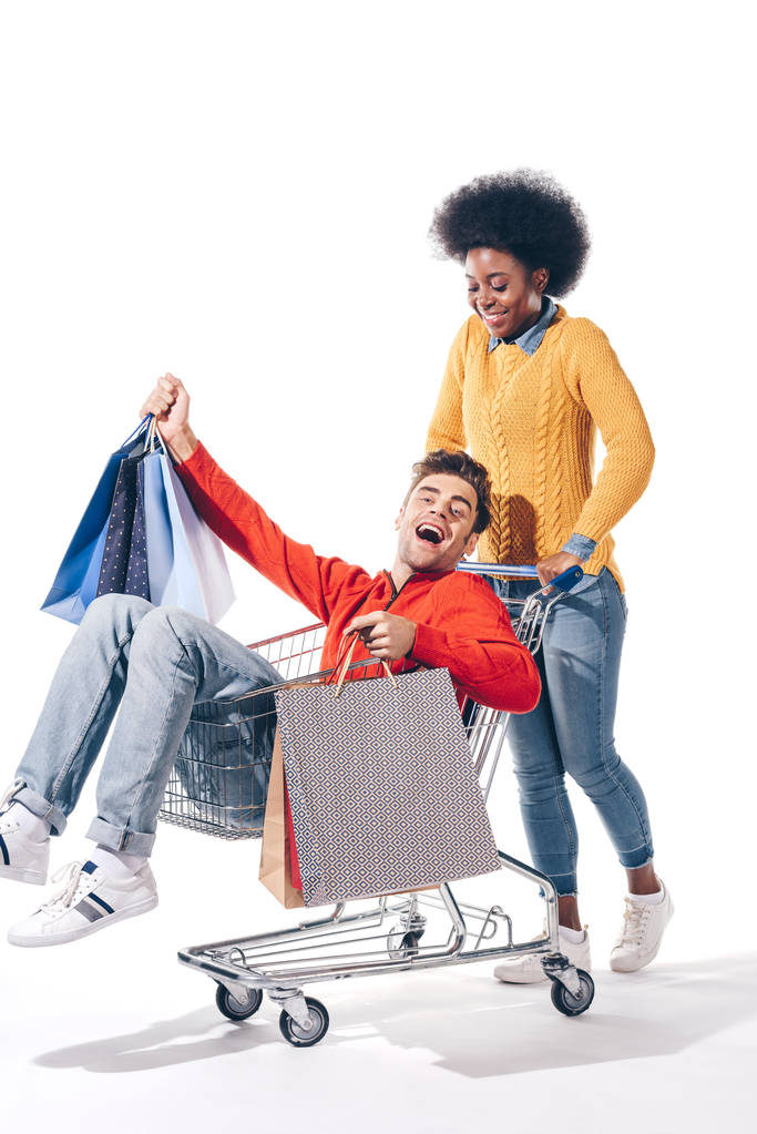 joven pareja multicultural divirtiéndose en carrito de la compra con bolsas de compras, aislado en blanco
 - Foto, Imagen