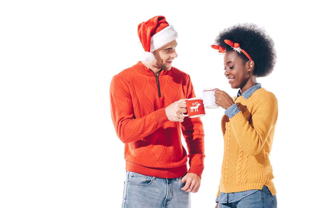 Lächelndes multikulturelles Paar mit Weihnachtsmann und Hirschhörnern in Kaffeetassen, isoliert auf weiß  - Foto, Bild