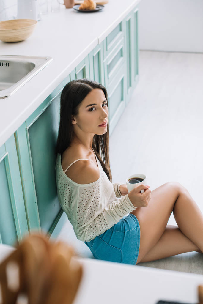 chica atractiva con taza de café sentado en el suelo en la cocina
 - Foto, Imagen