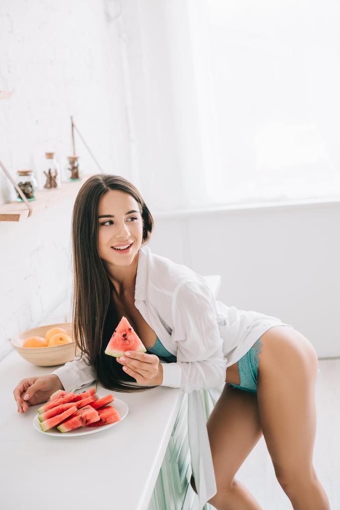 attractive happy girl in lingerie and white shirt eating watermelon in kitchen - Photo, Image