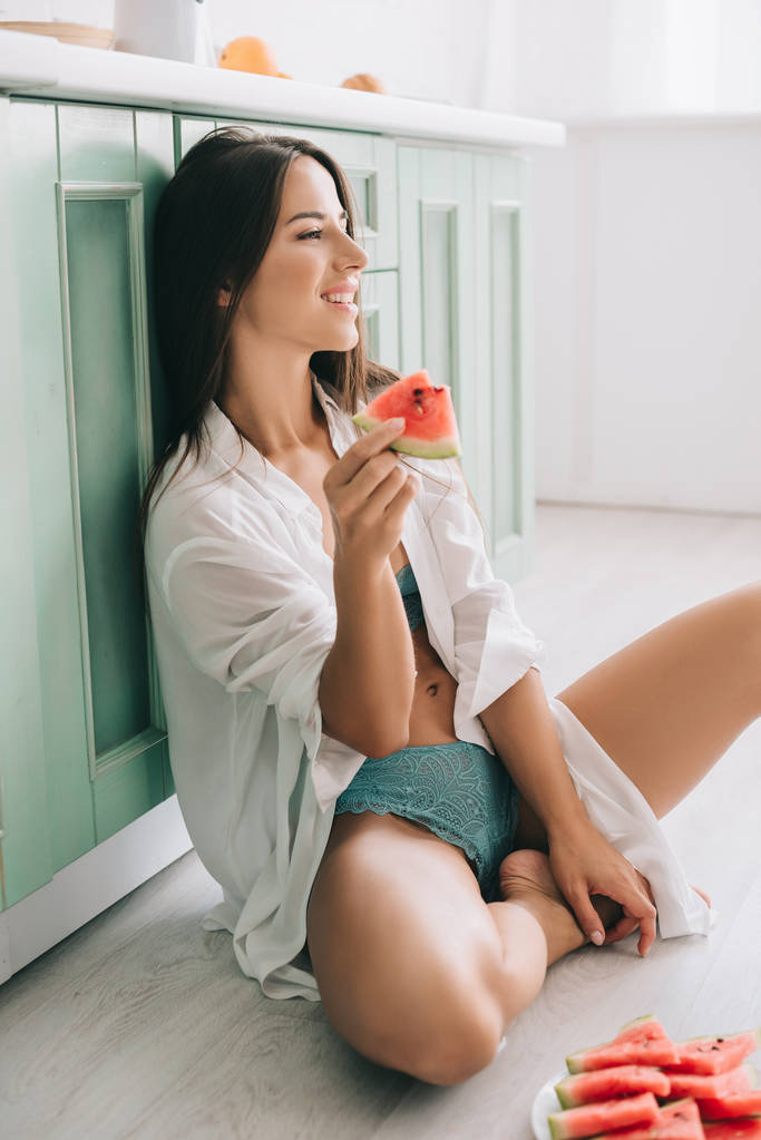 aantrekkelijke vrolijke vrouw in lingerie en wit shirt eten watermeloen op de vloer in de keuken - Foto, afbeelding