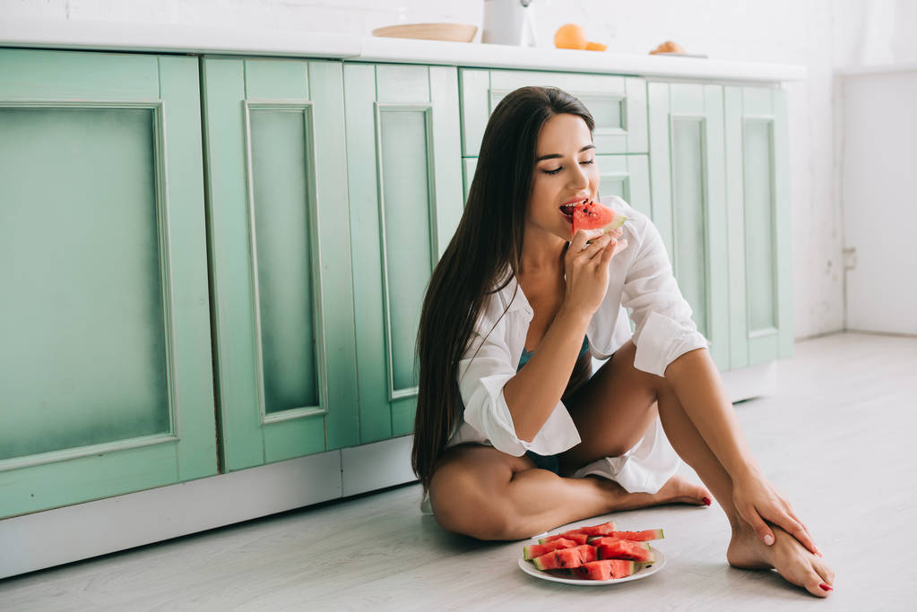 menina sexy em lingerie e camisa branca comendo pedaços de melancia no chão na cozinha
 - Foto, Imagem