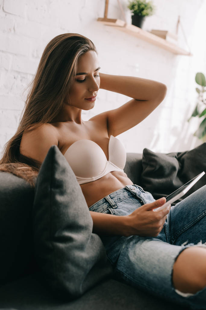 atrakcyjna dziewczyna w białym staniku i dżinsach za pomocą tabletu cyfrowego na kanapie w salonie - Zdjęcie, obraz