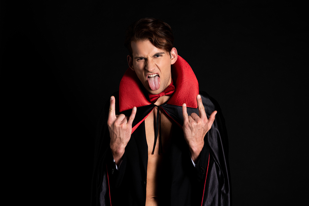 mężczyzna w halloween kostium wystający język i pokazując znak rocka izolowane na czarny  - Zdjęcie, obraz