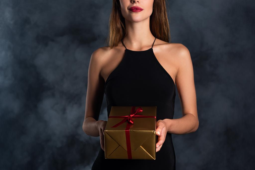 Ausgeschnittene Ansicht einer Frau mit Hörnern, die Geschenk auf schwarz mit Rauch hält  - Foto, Bild