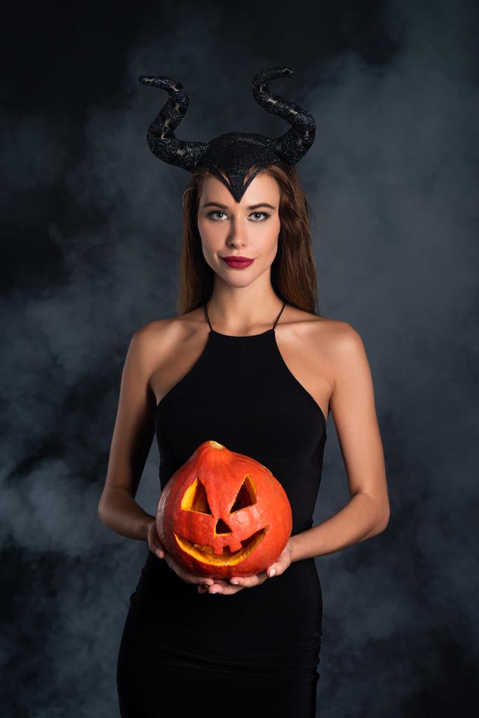 mulher atraente com chifres segurando abóbora halloween em preto com fumaça
  - Foto, Imagem