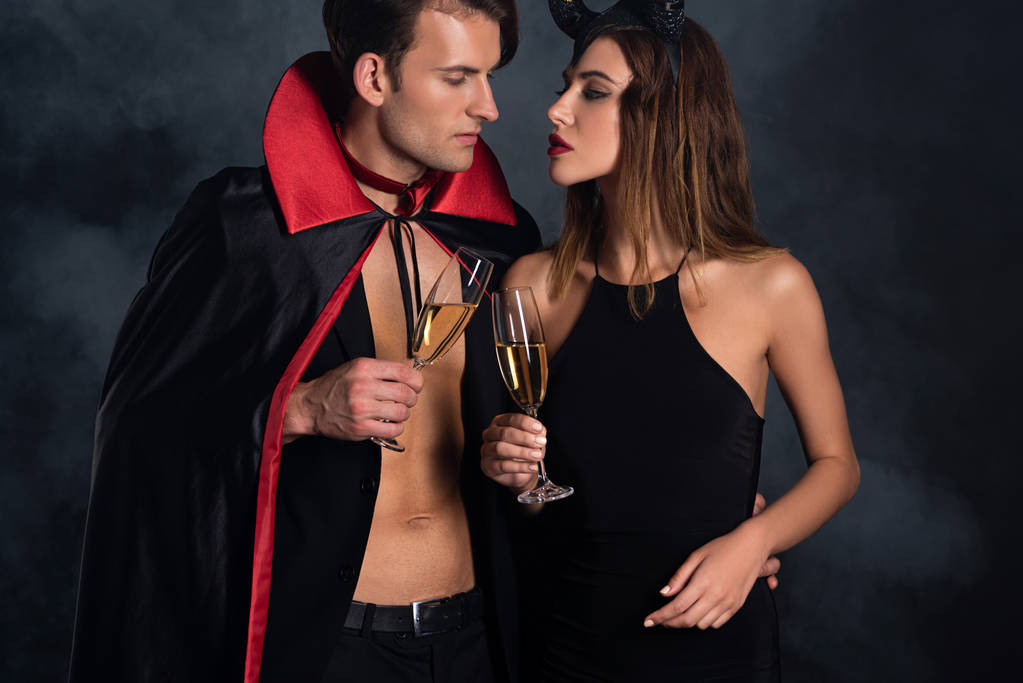 knappe man in mantel en vrouw met horens klinkende champagne bril op zwart met rook  - Foto, afbeelding