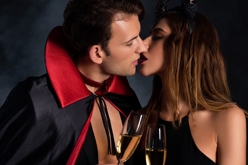 handsome man in cloak and woman with horns kissing and holding champagne glasses on black with smoke  - Photo, Image