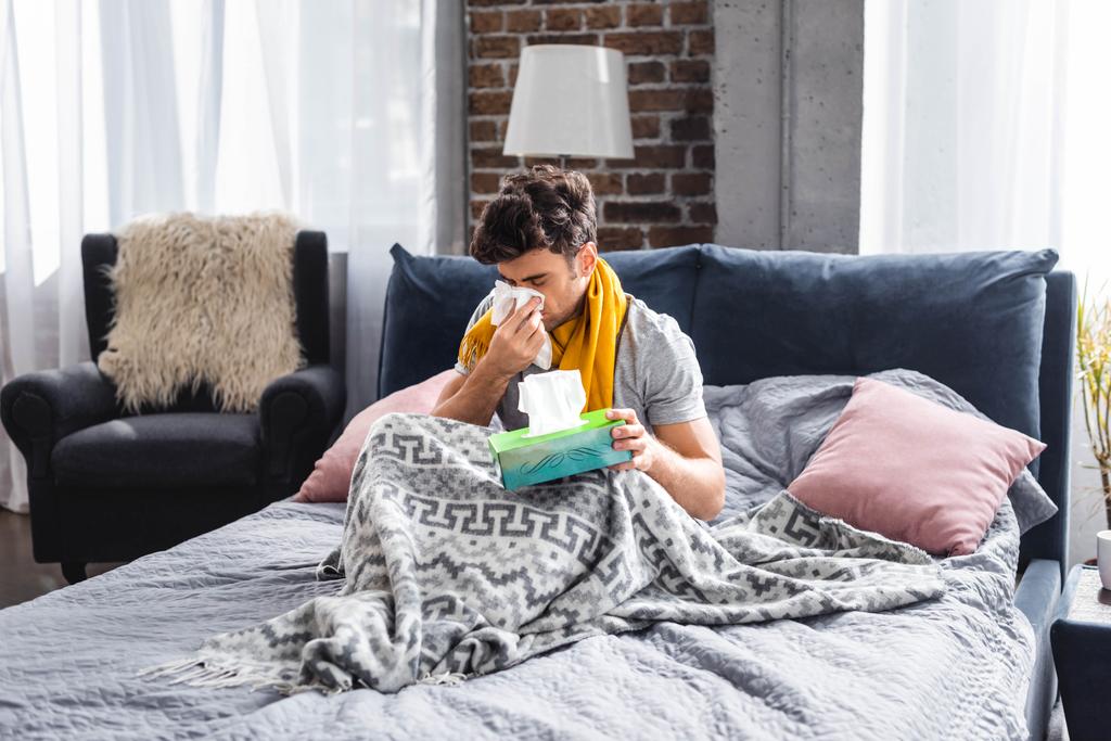 zieke man in sjaal niezen en houden servet in bed  - Foto, afbeelding