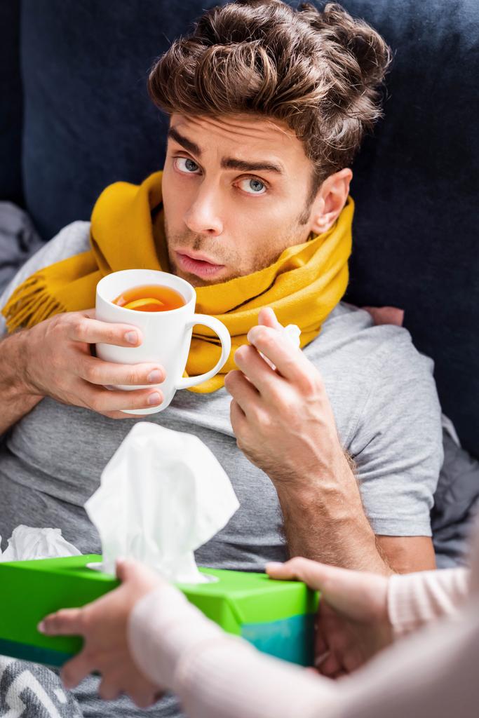 cropped view of girlfriend giving box with napkins to sick boyfriend with cup of tea  - Photo, Image