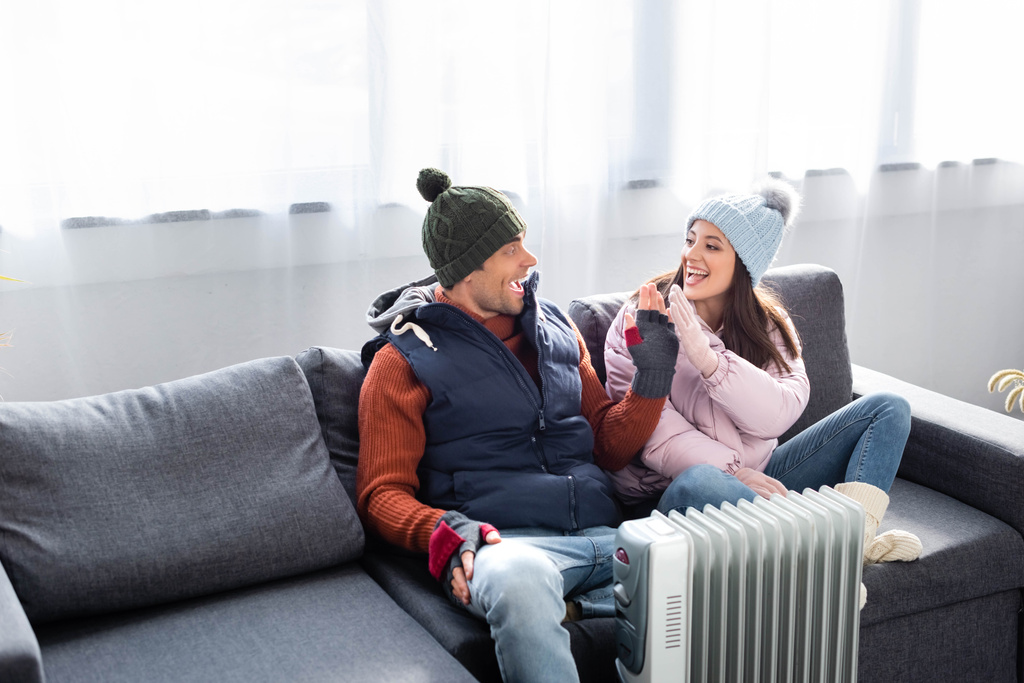 namorada atraente e namorado em roupa de inverno aquecendo perto do aquecedor e dando alta cinco
  - Foto, Imagem