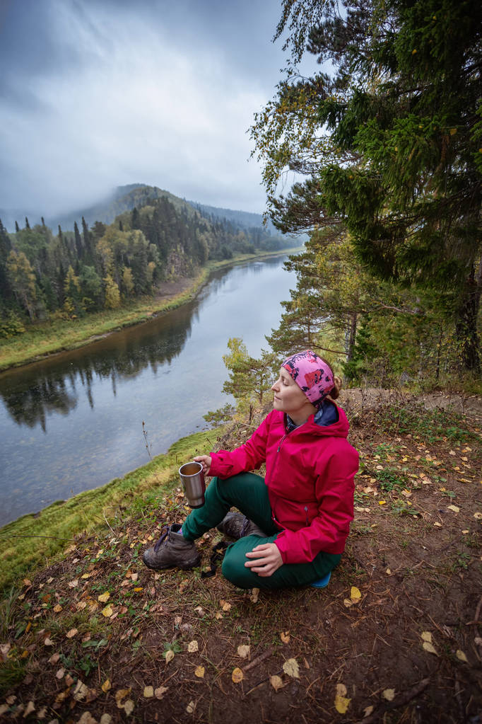 秋の風景。若い女性の観光客は、川のほとりにお茶を飲みながら座っている。川を見て。秋のキャンプ - 写真・画像