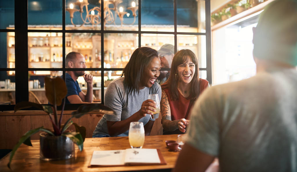 Gruppo sorridente di giovani amici seduti insieme a un tavolo in un bar nel pomeriggio ridendo e parlando di drink
 - Foto, immagini