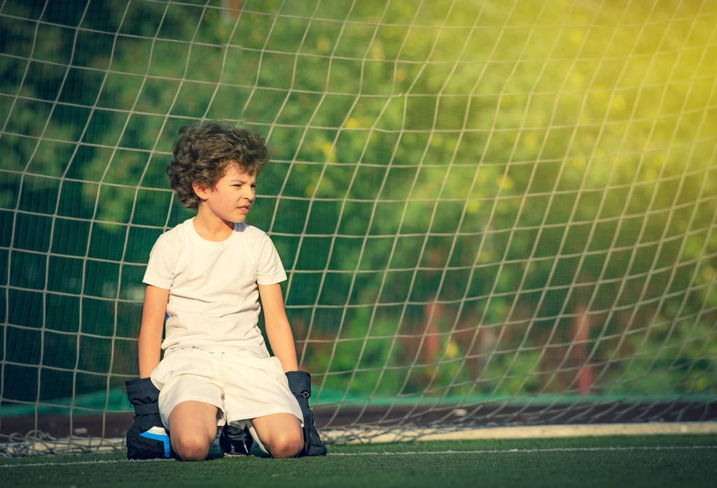 Düşünceli küçük çocuk kale direğindeki yeşil çimlerde oturan genç bir kalecinin kalesinde. Stadyumda futbol takımının erkek kalecisi. Spor kavramı. Kopya alanı - Fotoğraf, Görsel