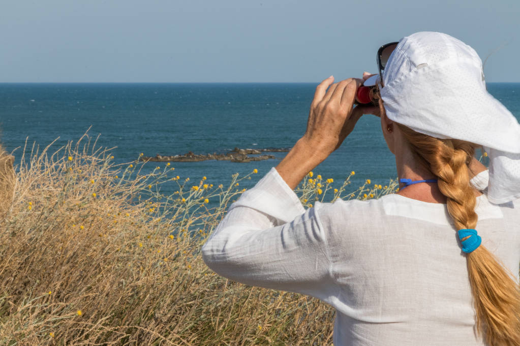 Beyaz bluzlu ve beyaz şapkalı yetişkin bir kadın, deniz kıyısında duruyor ve dürbünle bakıyor.. - Fotoğraf, Görsel