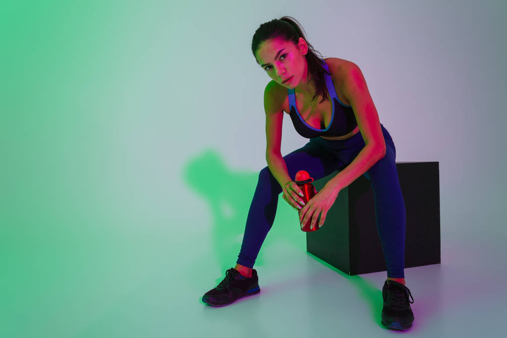 Serious young fitness woman posing isolated with led flash lights holding bottle with water have a rest. - Photo, Image