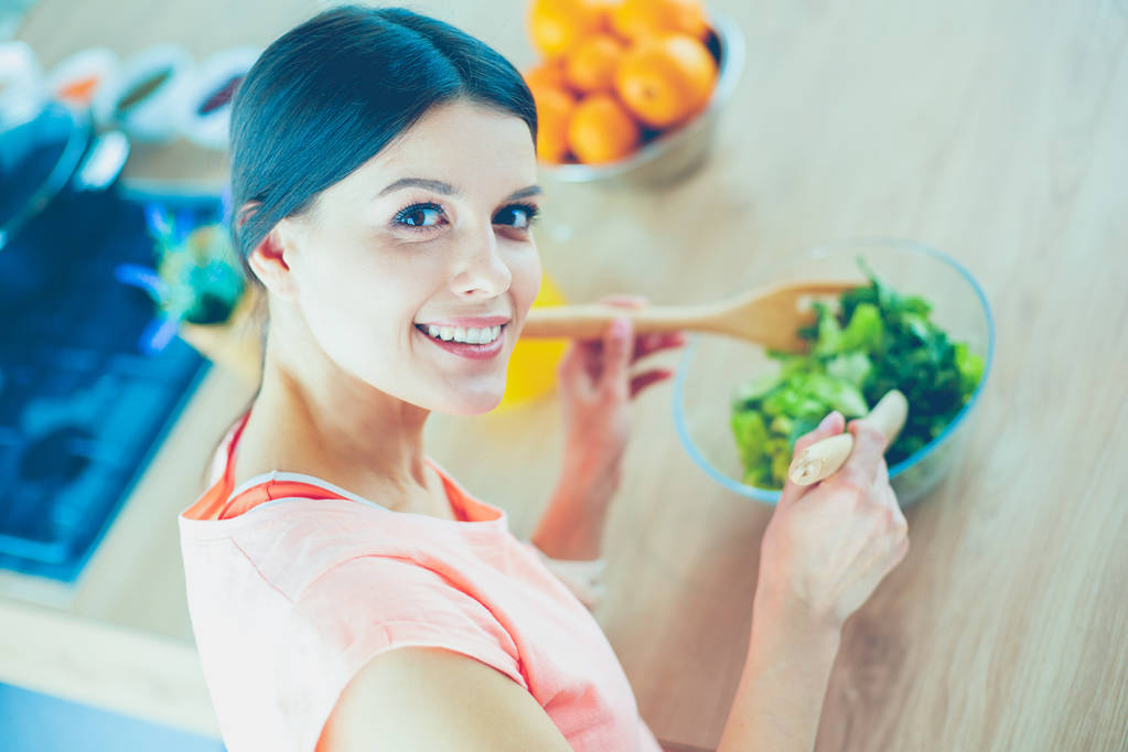 Улыбающаяся молодая женщина смешивает свежий салат на кухне. - Фото, изображение