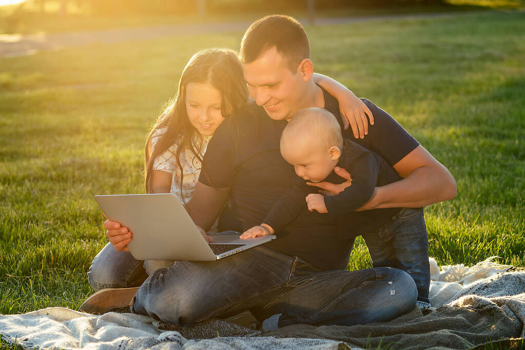 mały chłopiec wraz z ojcem i piękną córką bawią się i rozwijają patrząc na laptopa na kocu w parku w lecie - Zdjęcie, obraz