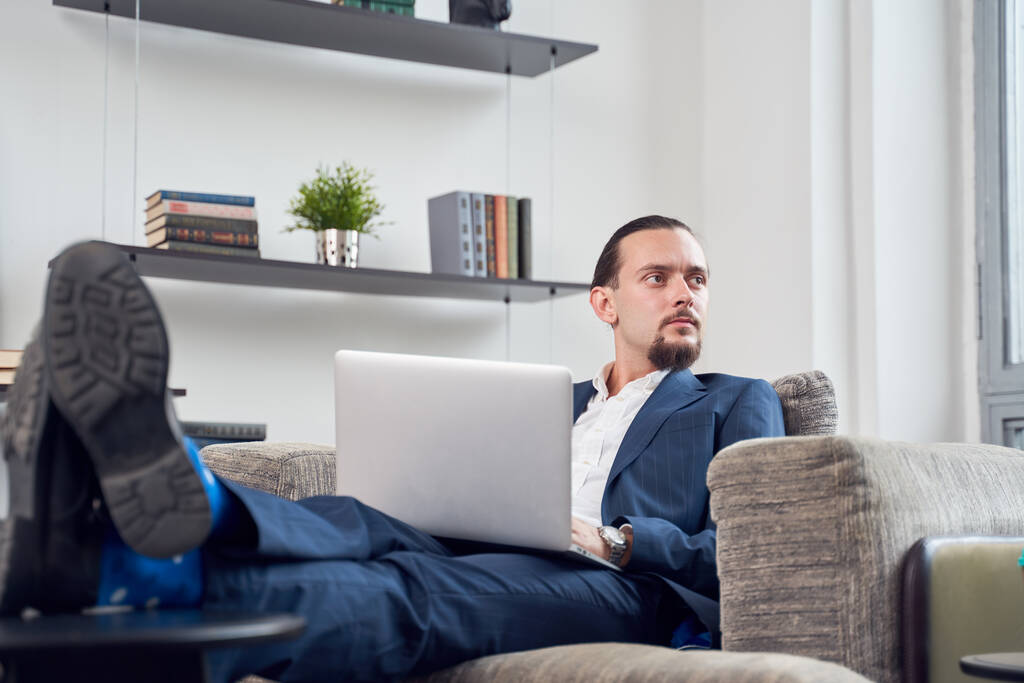 Εικόνα σοβαρού επιχειρηματία που κοιτάζει πλευρά με φορητό υπολογιστή κάθεται σε καναπέ στο δωμάτιο. - Φωτογραφία, εικόνα
