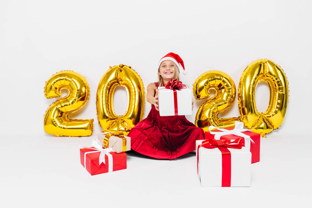 Счастливая маленькая девочка в шляпе Санта-Клауса, стильный малыш, делающий рождественский подарок, глядя на камеру, сидящую на белом фоне с золотыми цифрами 2020
 - Фото, изображение