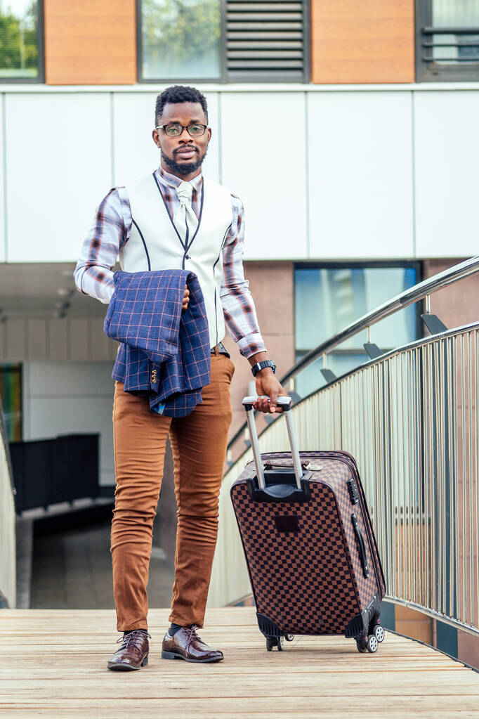 Başarılı ve iyi giyimli Afro-Amerikan iş adamının tam boy portresi. Havaalanında tekerlekli çantasıyla yürüyor. Tekerlekli büyük bir bavul. - Fotoğraf, Görsel