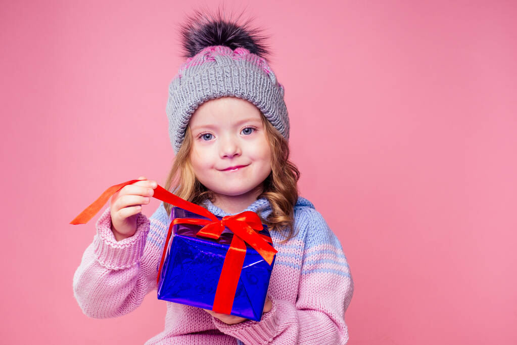 Feliz niña peinado rizado rubia sonriente en sombrero gris de punto con caja de regalo de Navidad con lazo en el estudio de fondo rosa. nuevo Año presente en las manos de una niña pidiendo un copyspace deseo
 - Foto, imagen