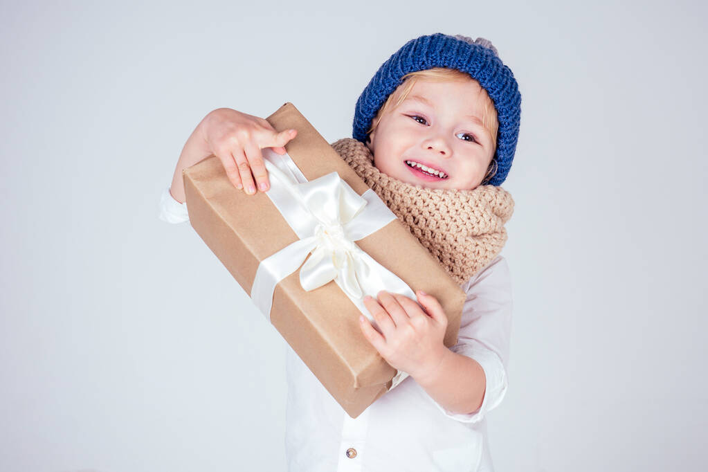 Happy little smiling blonde curly hairstyle boy in a knitted blue hat with christmas gift box with bow on white background in studio. new Year present in the hands of a male child - Photo, Image