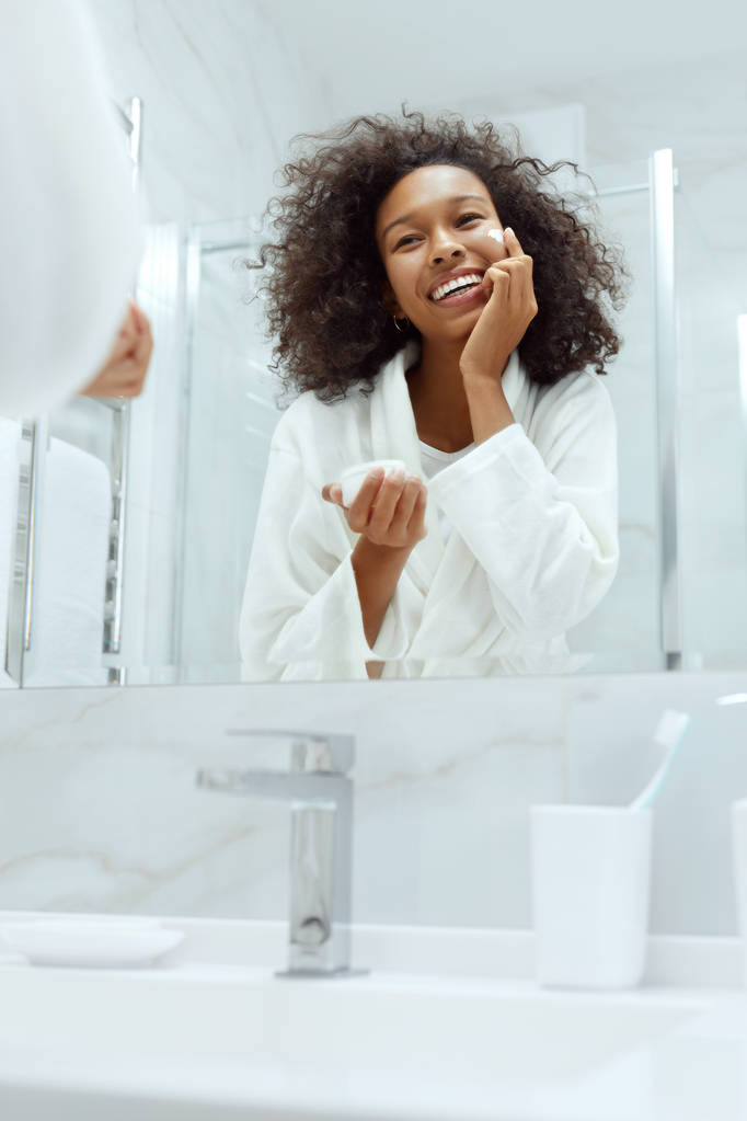 Péče o pleť. Žena aplikující krém na obličej při pohledu v koupelně zrcadlo - Fotografie, Obrázek
